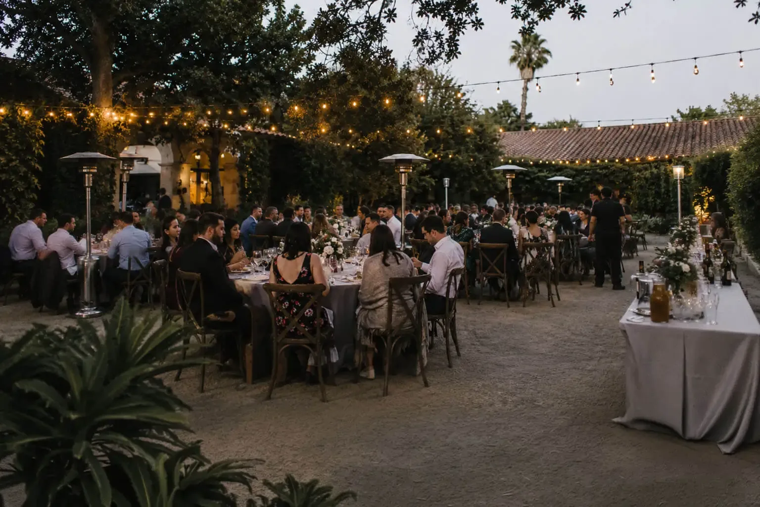 Wedding catering in Oakland, CA by Oakland Catering Company — staffed courtyard reception dinner service, rentals, floral tablescapes, cake service and bar service for weddings in Oakland, Berkeley, Alameda, Emeryville, Piedmont and San Leandro Twilight garden courtyard wedding reception in Oakland, CA — guests dining at round and long tables with white floral centerpieces under bistro string lights and patio heaters, beside a tile-roofed villa and a linen-draped bar with glassware and wine