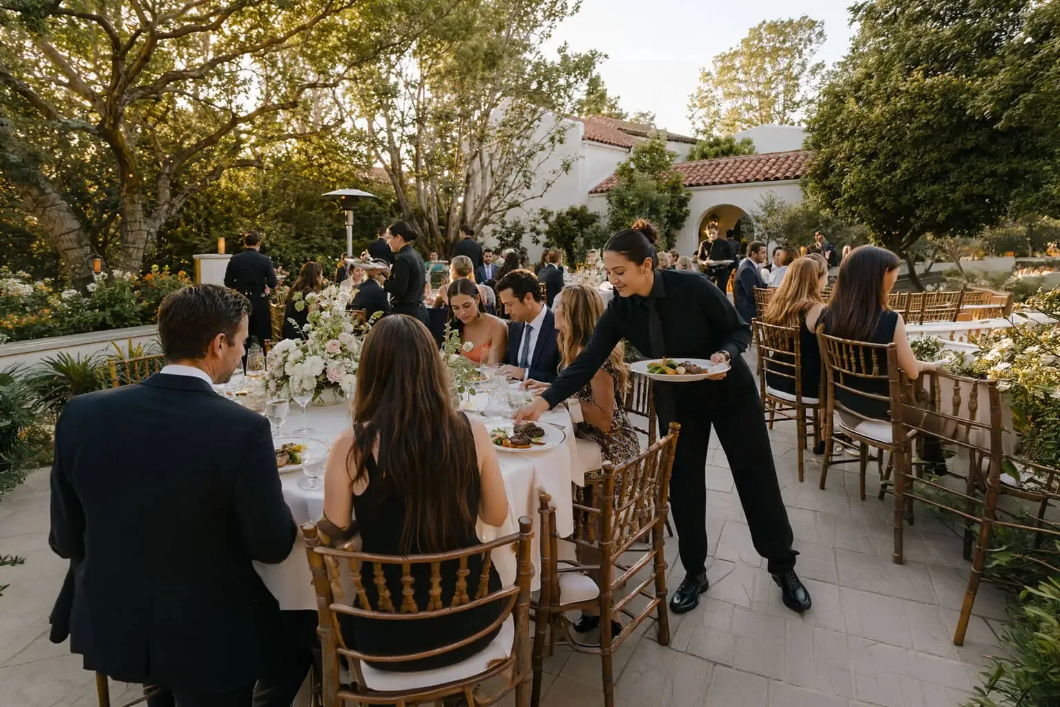 Wedding catering in Palo Alto, CA by Palo Alto Catering Company — plated reception service with uniformed staff, rentals, floral tablescapes, cake service and bar service for weddings in Palo Alto, Menlo Park, Stanford, Mountain View, Los Altos and Redwood City Golden-hour garden wedding reception in Palo Alto, CA — a black-clad catering attendant sets down a plated entrée at a linen table with blush and ivory floral centerpieces, guests seated in gold chiavari chairs beside a Spanish tile-roofed estate
