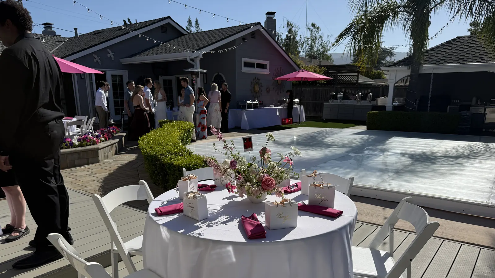 Event rentals in Piedmont and Oakland, CA by Oakland Catering Company — gender reveal party round tables, floor-length linens, napkins, place settings, white folding chairs, pink market umbrellas, patio heaters and chafing dish buffet rentals with delivery and setup for backyard events in Oakland, Piedmont, Berkeley, Alameda, Emeryville and San Leandro Gender reveal party event rentals in a Piedmont, CA residential backyard — round table draped in a floor-length white linen with hot pink napkins, gold-lettered thank-you favor boxes and a pink and white floral centerpiece, surrounded by white folding chairs on a deck beside a covered pool, with pink market umbrellas, patio heaters and linen-draped buffet tables of silver chafing dishes