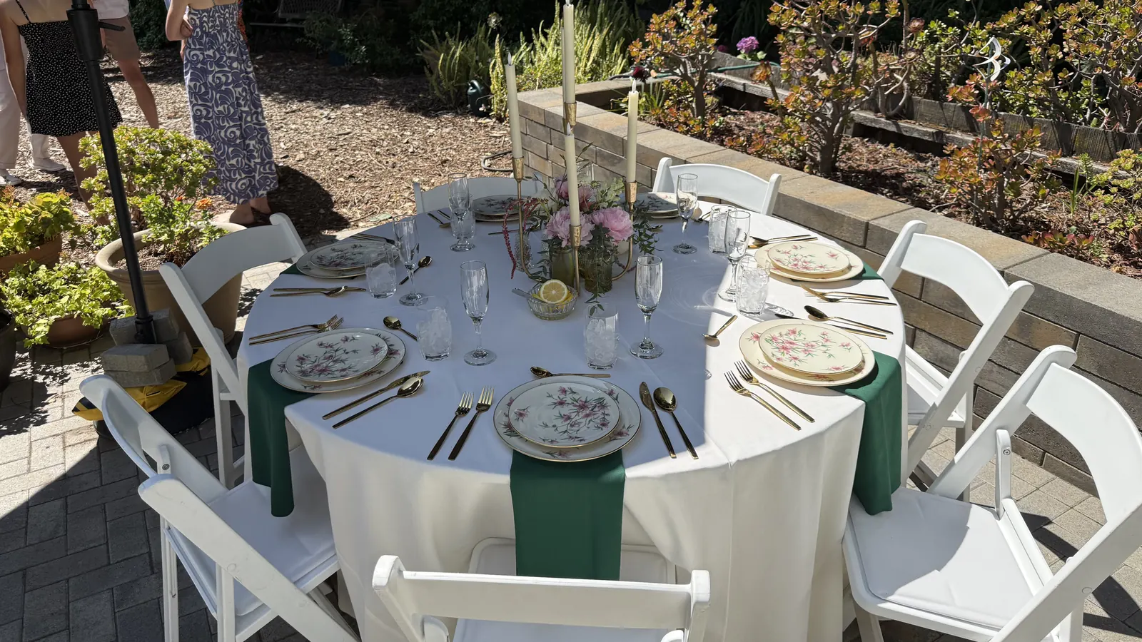Event rentals in Palo Alto, CA by Palo Alto Catering Company — engagement party table, linen, chair, china, glassware and place setting rentals with delivery and setup for backyard and at-home events in Palo Alto, Menlo Park, Stanford, Mountain View, Los Altos and Redwood City Engagement party event rentals in a Palo Alto residential backyard — a round table draped in white linen with emerald green runners, vintage floral china place settings, gold flatware, crystal stemware, taper candles and blush rose centerpieces, surrounded by white folding garden chairs on a paver patio