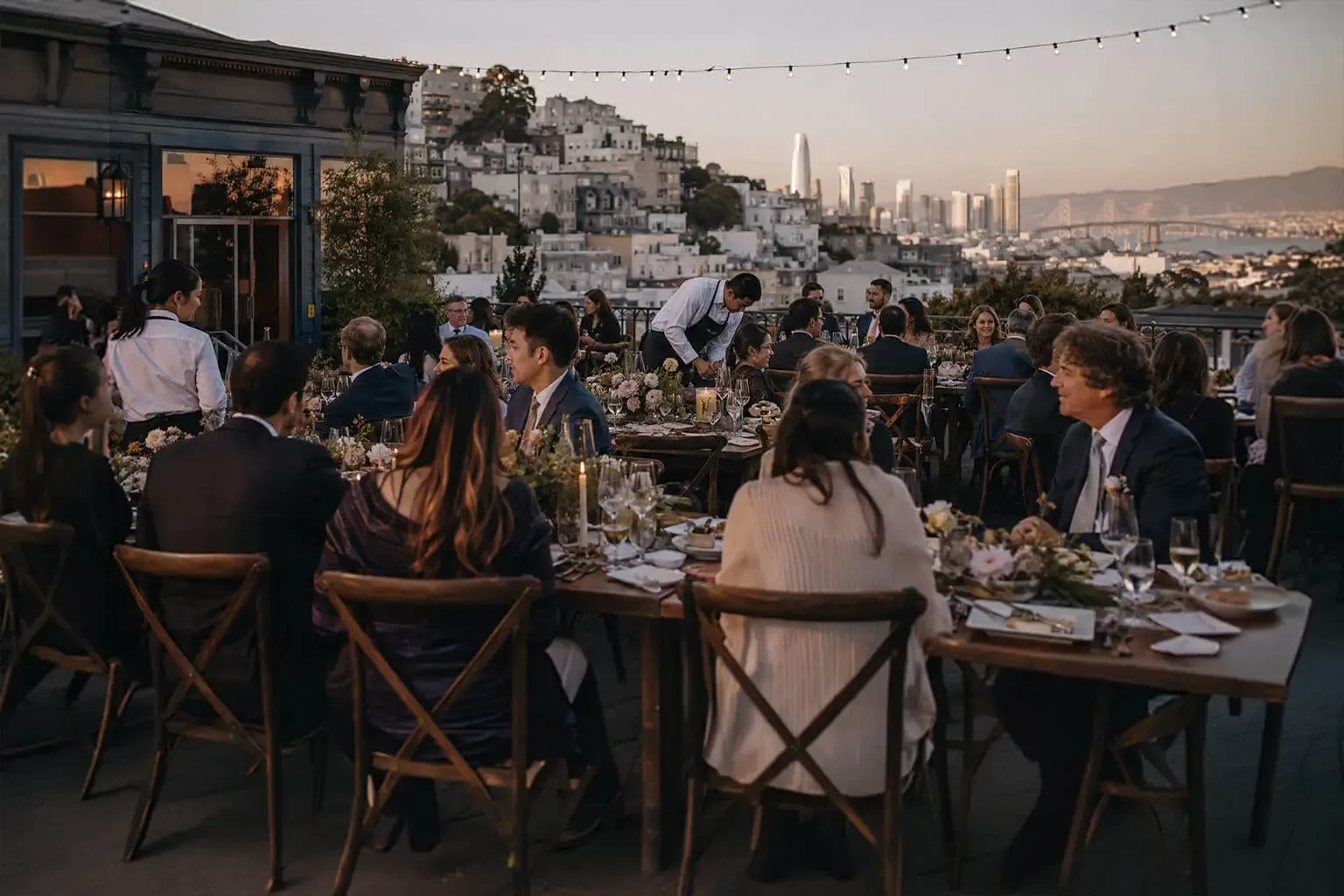 Winslow's Catering San Francisco — wedding catering on a hilltop rooftop terrace above the city: attendants passing plated courses at candlelit farm tables with garden-rose arrangements, Victorian rooftops, the downtown skyline and the Bay Bridge in view, serving San Francisco, Daly City, Brisbane, Colma and South San Francisco Sunset rooftop terrace wedding reception in San Francisco, CA, with guests dining at long wooden tables under cafe lights and the city skyline and Bay Bridge beyond — wedding catering by Winslow's Catering San Francisco