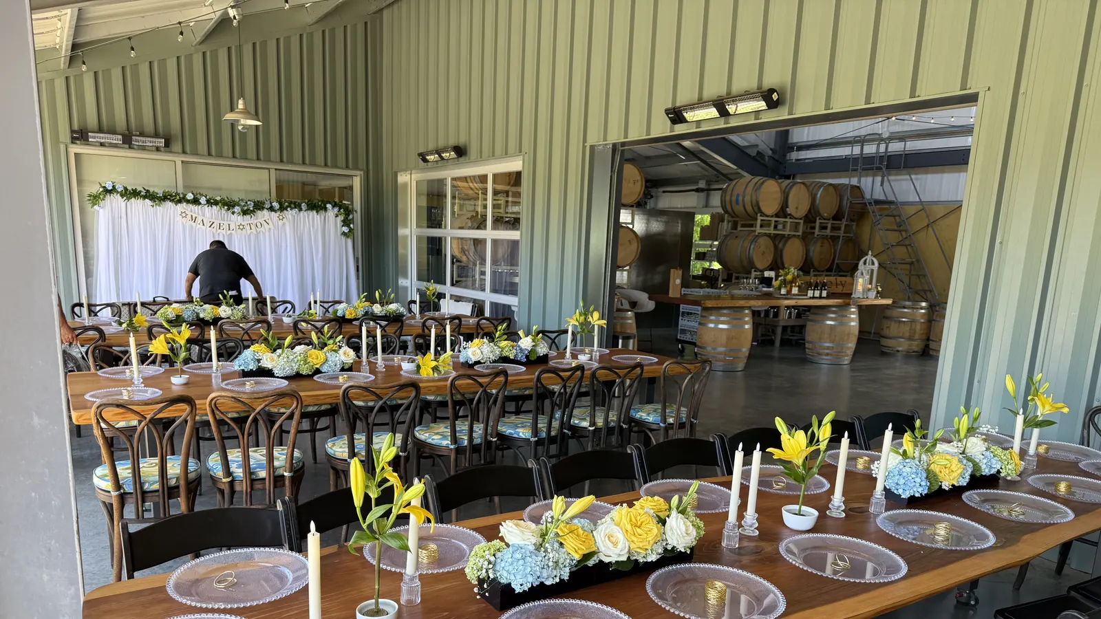 Event rentals in San Francisco, CA by SF Catering Company — 16th birthday party farm tables, chairs, linens, charger plates, glassware, candles and full place setting rentals with delivery and setup for venues in San Francisco, Daly City, Brisbane and South San Francisco Sweet 16 birthday party event rentals at a San Francisco venue — long wooden farm tables set with beaded glass charger plates, gold napkin rings, ivory taper candles in glass holders and blue hydrangea, yellow rose and lily centerpieces, lined with dark bentwood and black folding chairs beneath a 'Mazel Tov' floral backdrop