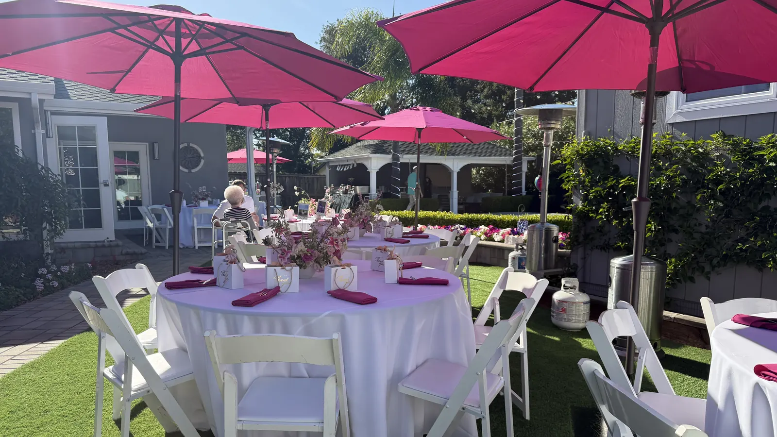 Event rentals in Lafayette and Walnut Creek, CA by Walnut Creek Catering Company — surprise engagement party round tables, floor-length linens, napkins, white folding chairs, market umbrellas and patio heater rentals with delivery and setup for at-home events in Walnut Creek, Lafayette, Danville, Alamo, Orinda, Pleasant Hill and Concord Surprise engagement party event rentals in a Lafayette, CA backyard — round tables draped in white floor-length linens with fuchsia napkins, gold-lettered thank-you favor bags and dusty rose floral centerpieces, surrounded by white folding garden chairs under hot pink market umbrellas with patio heaters and a white gazebo on the lawn