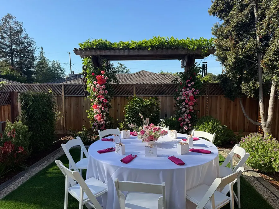 Rose arch in pink and red framing a round table with white linens and fuchsia napkins in a garden — custom floral installation styling for Tracy catering events